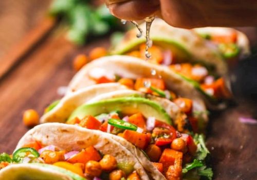 The image shows a hand squeezing lime over tacos filled with chickpeas, avocado, and vegetables, garnished with fresh herbs on a wooden board.