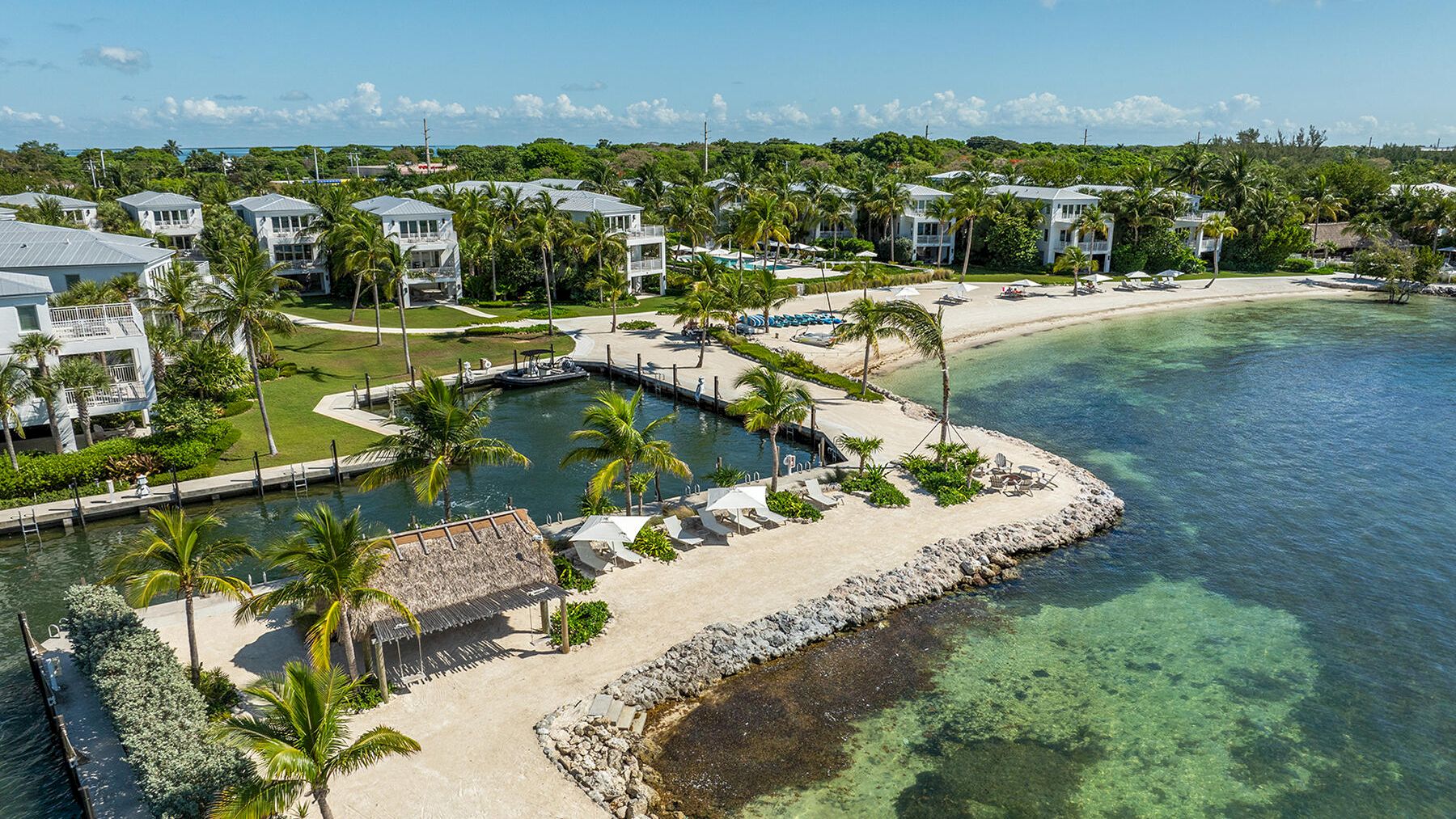 A picturesque beachfront resort with white buildings, lush greenery, palm trees, a sandy beach, and clear, calm ocean water covering rocks.