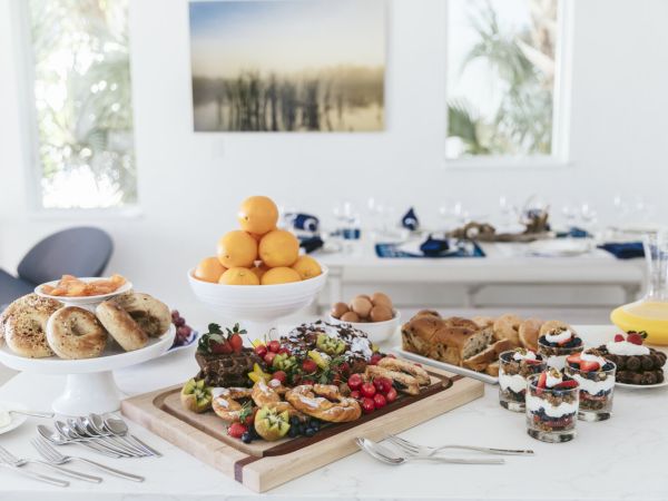 A table displaying a variety of breakfast foods, including bagels, fruit parfaits, pastries, fresh fruit, and juice, set in a bright dining room.
