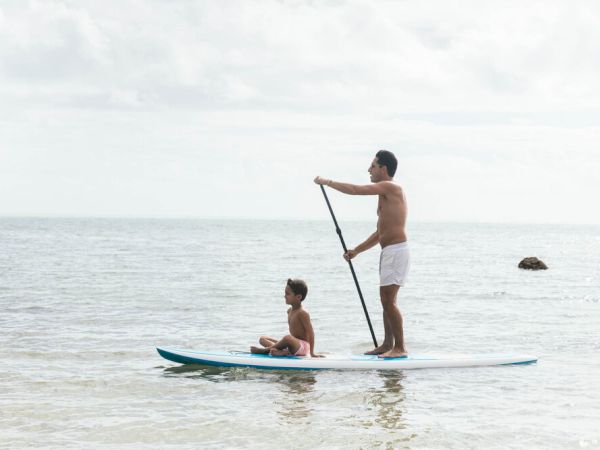 A person is stand-up paddleboarding with a child sitting in front on a calm ocean water. They both appear to be enjoying a leisurely time.