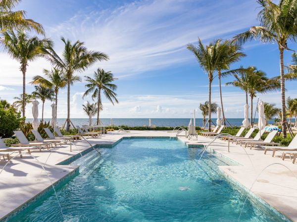 A luxurious pool area with sun loungers and palm trees, overlooking a serene ocean view under a clear blue sky with some wispy clouds.