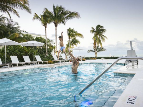 A person lifts a child in a swimming pool surrounded by lounge chairs and palm trees, under a clear sky.
