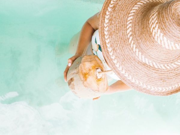 A person wearing a wide-brimmed hat stands or floats in clear water while holding and drinking from a coconut.
