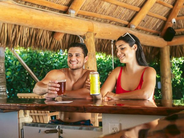 A man and woman sit at a tiki bar, smiling and enjoying drinks with a wooden roof and lush green background.