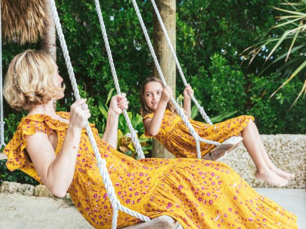 Two people in yellow dresses are sitting on swings under a rustic pergola, surrounded by lush greenery.