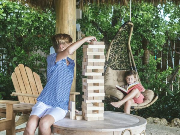 A person plays Jenga on a table outdoors, while another sits in a hanging chair. A bag is on the ground.