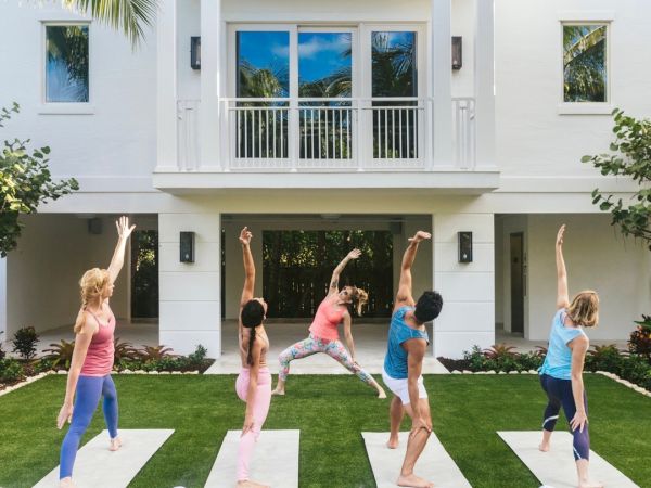 People practicing yoga on mats in a garden in front of a white building, surrounded by trees, engaging in different poses.