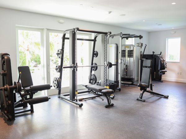 A home gym with various exercise equipment, including weight machines, benches, and a Smith machine, in a bright room with windows.