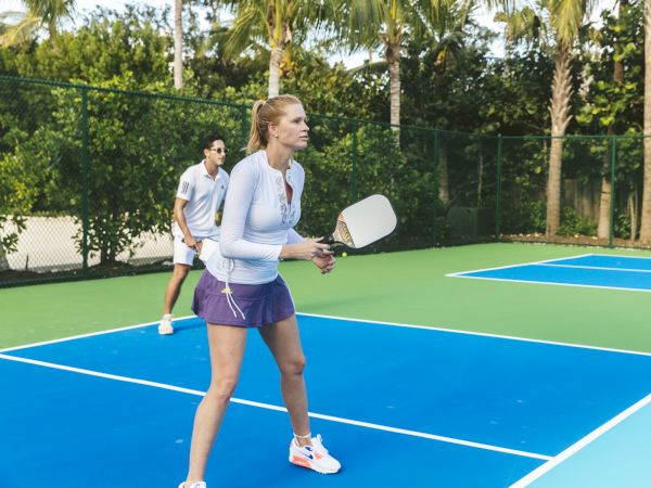 Two people are playing pickleball on an outdoor court surrounded by trees and a fence. The woman is in the foreground, preparing to hit the ball.