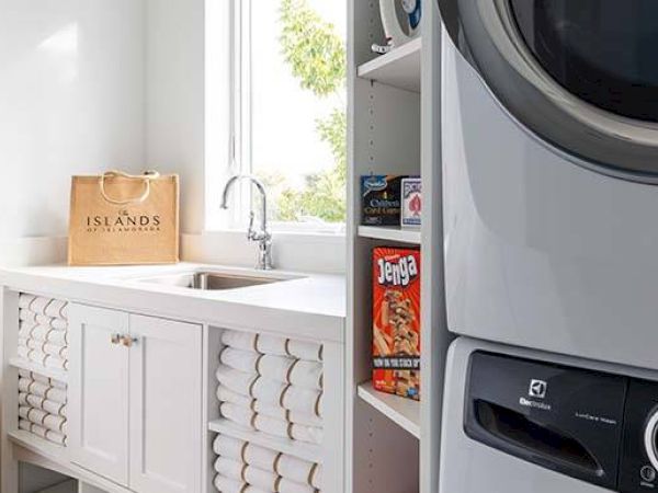 A laundry room with a sink, stacked washer/dryer, cabinet with neatly rolled towels, and shelves with board games and detergent.