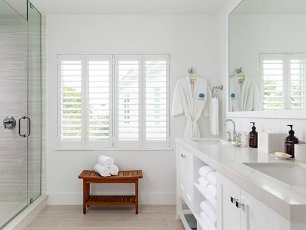 A modern bathroom with a glass-enclosed shower, double sink counter, robes on hooks, and folded towels on a wooden bench by a window.
