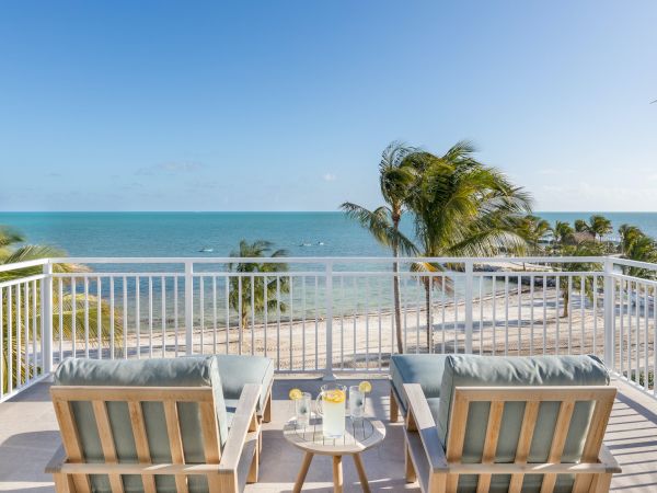 A scenic oceanview balcony with two lounge chairs, a table with drinks, and palm trees, overlooking a sandy beach under a clear blue sky.