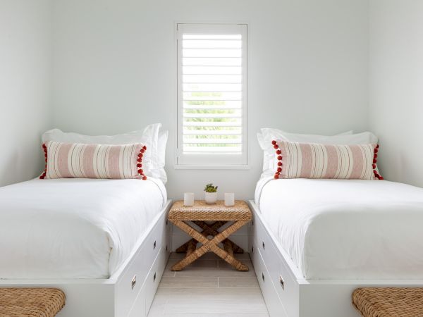 A minimalist bedroom with two single beds, a central wicker nightstand, white linens, striped pillows, and a small window with shutters.