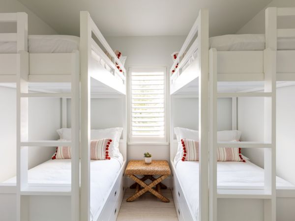 The image shows a bedroom with two sets of white bunk beds, a window with shutters, and a small woven table in the center, creating a cozy atmosphere.