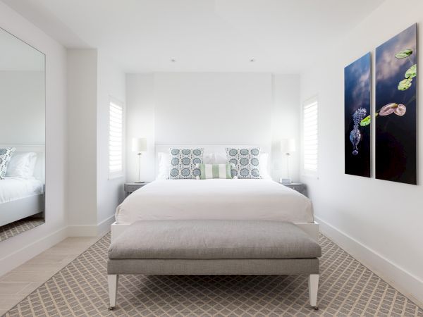 A modern bedroom with a bed, a bench at the foot, two side tables with lamps, a large mirror, and wall art featuring lily pads and flowers.