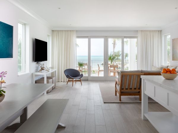 A bright, modern living room with white walls and furnishings, overlooking a balcony with a beach view, featuring a TV, sofa, and dining area.