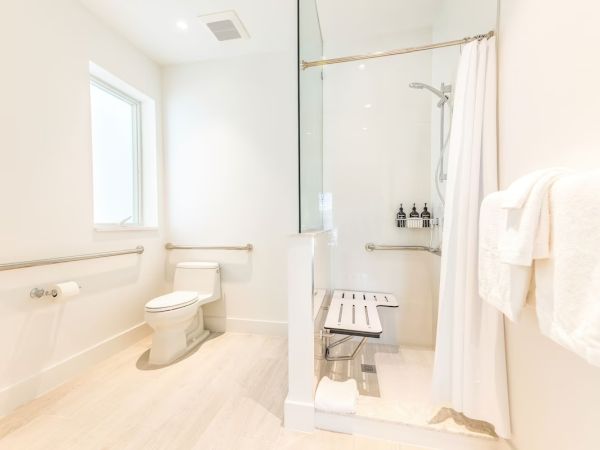 A clean, white bathroom featuring a toilet, shower area with a bench and grab bars, a towel rack with towels, and a window for natural light.