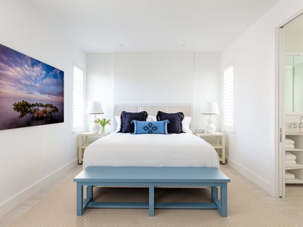A well-lit bedroom with a bed, blue bench, white bedding, and two nightstands with lamps. A colorful painting adorns the wall. A bathroom is adjacent.
