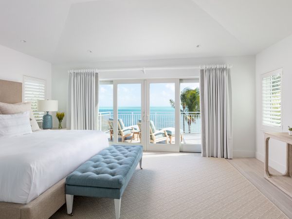 A bright bedroom with a large bed, blue bench, TV, and ocean view through glass doors leading to a balcony with seating.