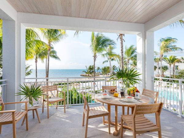 A patio with wooden furniture, breakfast items on the table, overlooking a pool, palm trees, and a beach with a clear blue sky in the background.