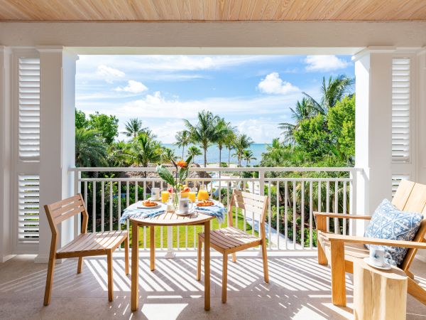 A balcony with a wooden table set for breakfast, overlooking a lush garden and the ocean, framed by palm trees and a bright, sunny sky.