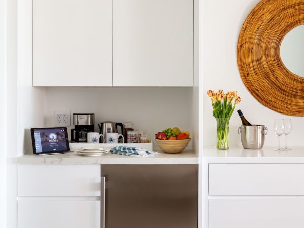 A modern kitchen corner with white cabinets, a stainless-steel fridge, a countertop with kitchen gadgets, flowers, a fruit bowl, and a round mirror.