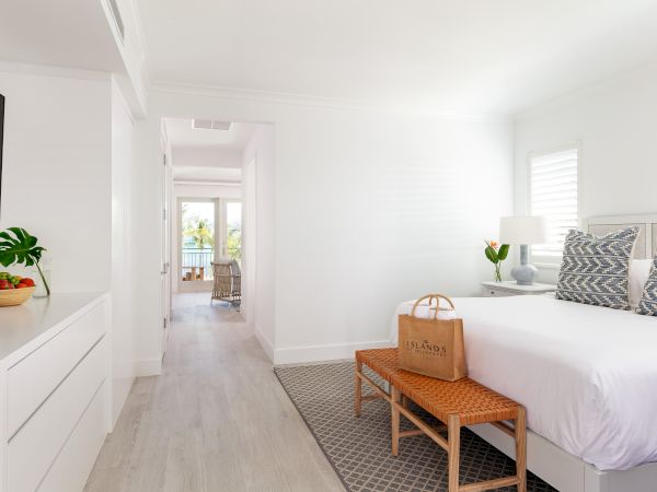 A bright bedroom with a bed, decorative pillows, plants, and a bag. There's an open hallway leading to a balcony with palm trees outside.