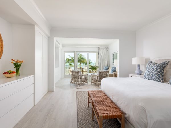 A bright, airy bedroom with a large bed, a sitting area, tropical decor, and a view of palm trees through sliding glass doors.