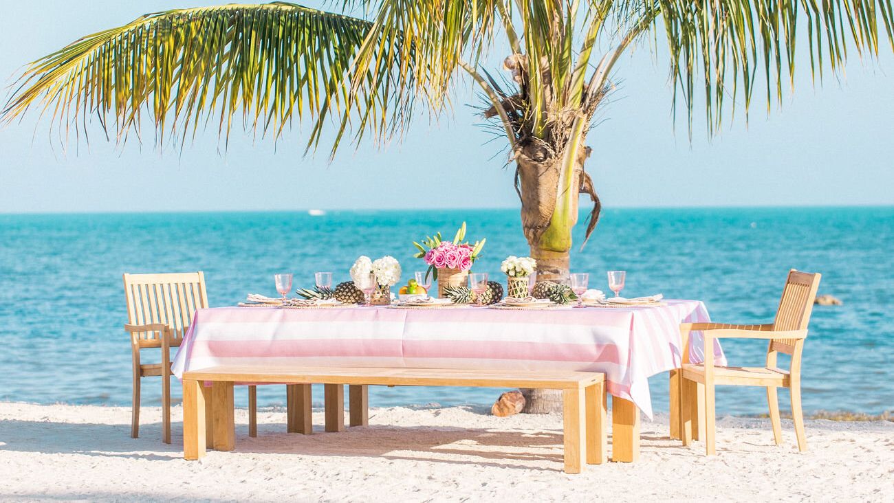 A beachside table with a pink tablecloth is set with flowers and dishes, shaded by a palm tree, overlooking the ocean.