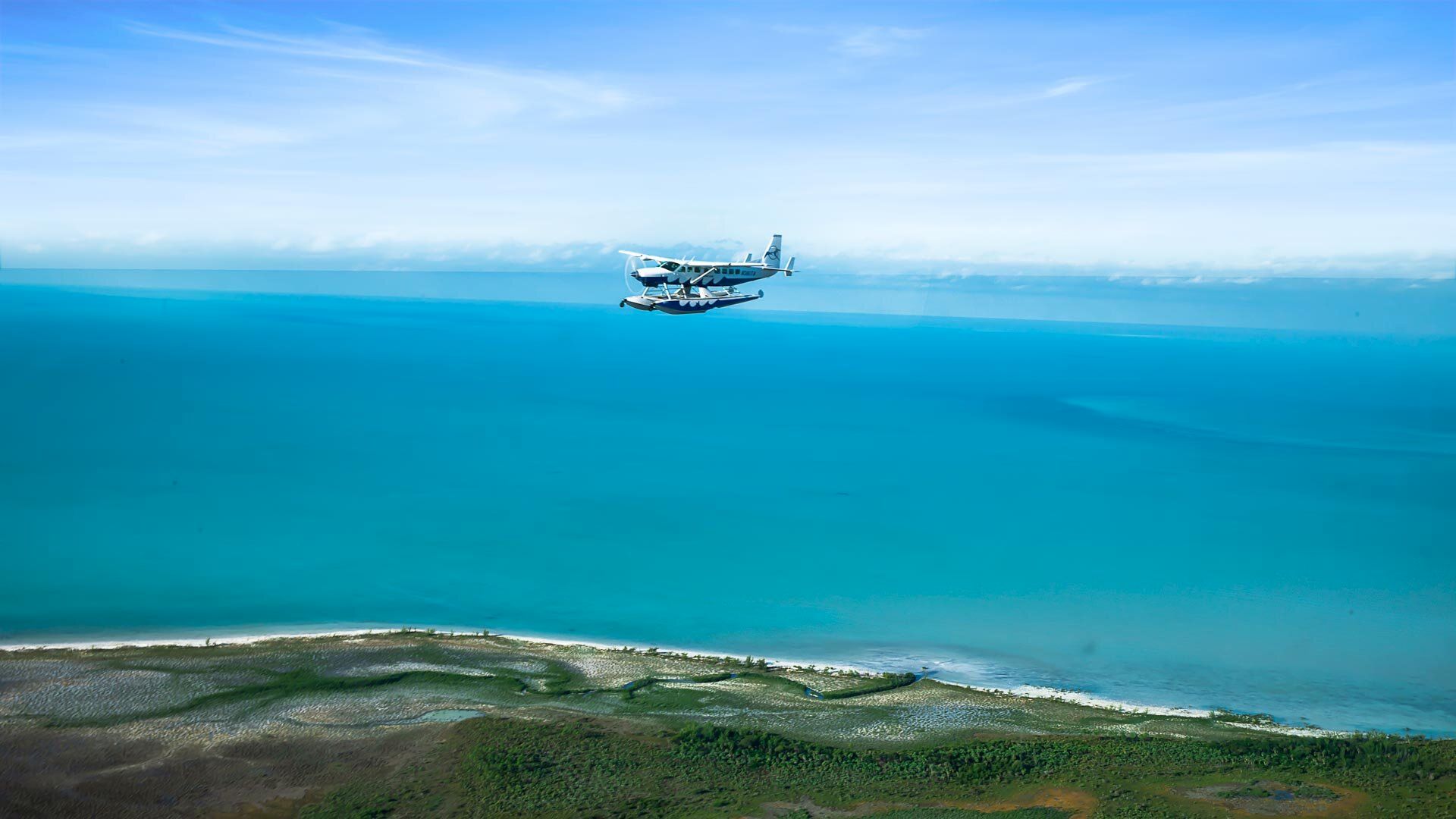 A small seaplane flies over a vast turquoise ocean with a green coastline visible below. The sky above is clear with some light clouds on the horizon.