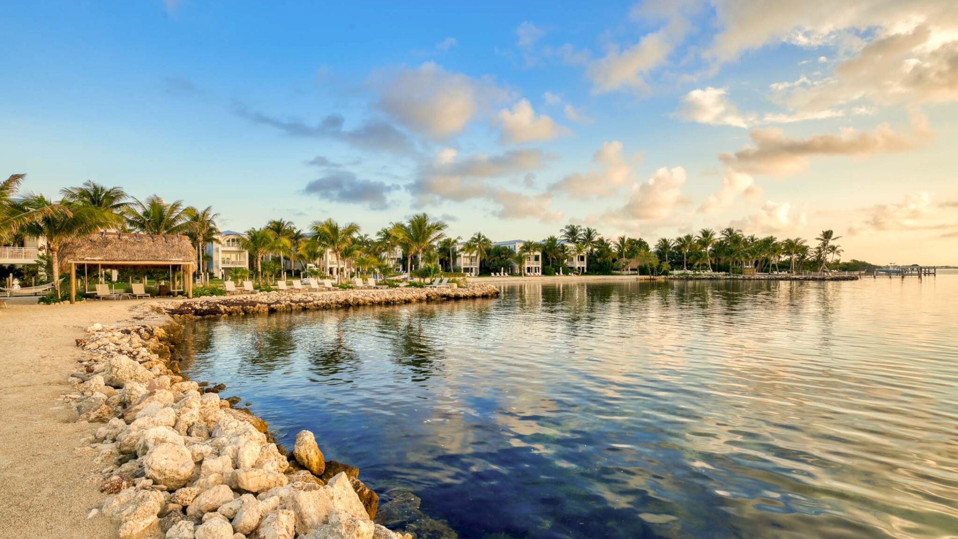 The image shows a scenic coastal view with a rocky shoreline, palm trees, and houses in the background under a blue sky with clouds at sunset.