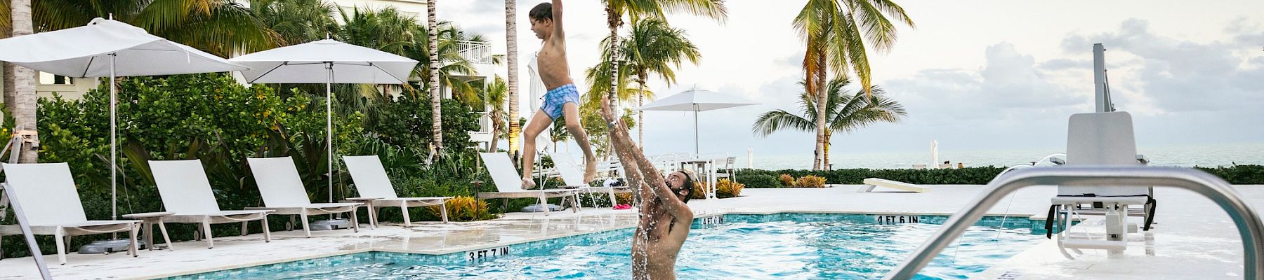 Two people jumping into a bright pool at a tropical resort with palm trees and lounge chairs.