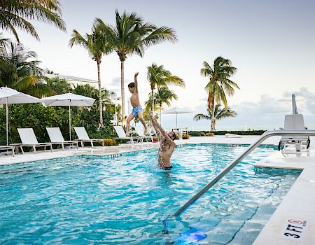 Two people jumping into a bright pool at a tropical resort with palm trees and lounge chairs.