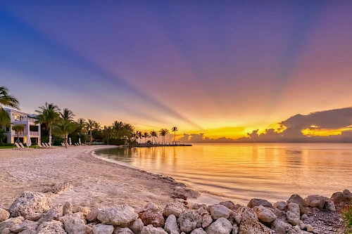 A tranquil beach sunset with orange-pink sky, gentle waves, palm trees, and rocky shoreline by calm water, a peaceful tropical retreat.