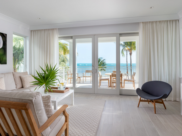 Bright beachside living room with white furniture, large windows, and an ocean view in the distance.