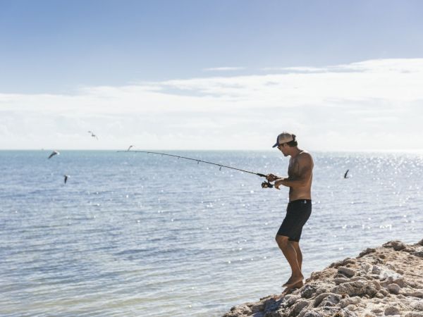 A person is standing on rocky terrain near the sea, fishing with a fishing rod, and wearing shorts and a cap.