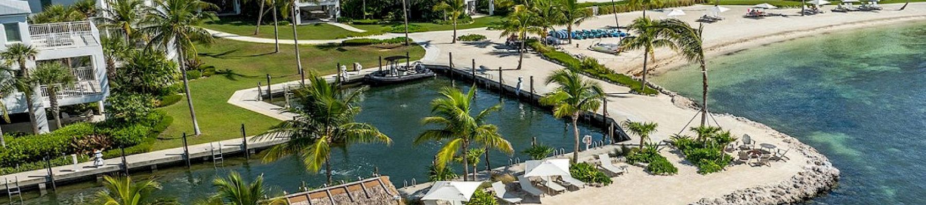 A tropical beachfront area with clear waters, palm trees, a small hut, and residential buildings in the background, under a blue sky.