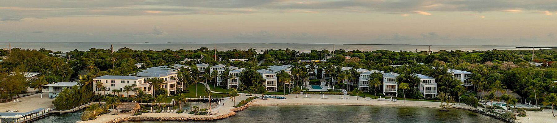The image displays a coastal area with a small beachfront neighborhood, lush greenery, and calm waters under a partly cloudy sky.