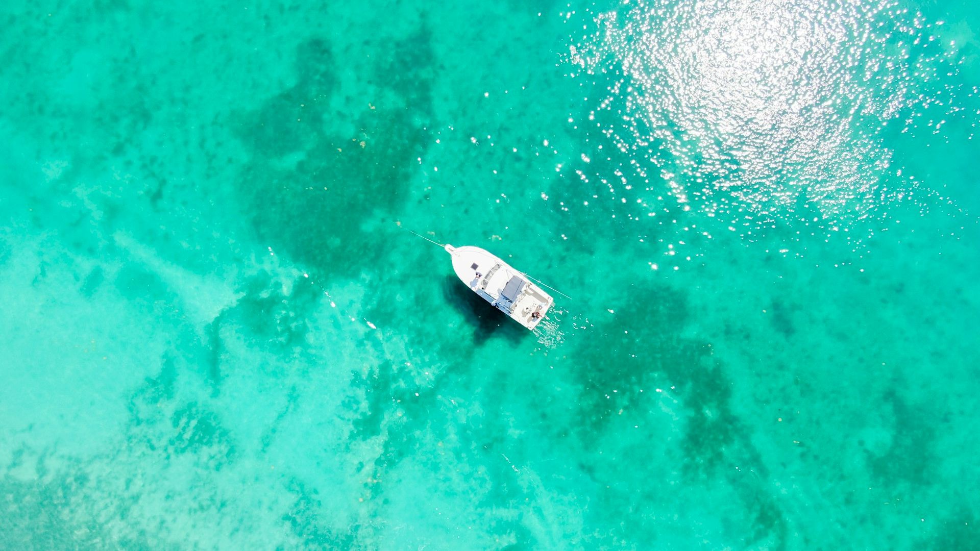 Aerial view of a boat floating on clear turquoise water, with sunlight creating a bright reflection on the surface.