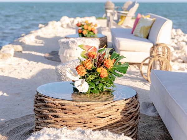 A seaside lounge area with stylish seating, cushions, and decorative flowers on woven tables overlooks a calm ocean.