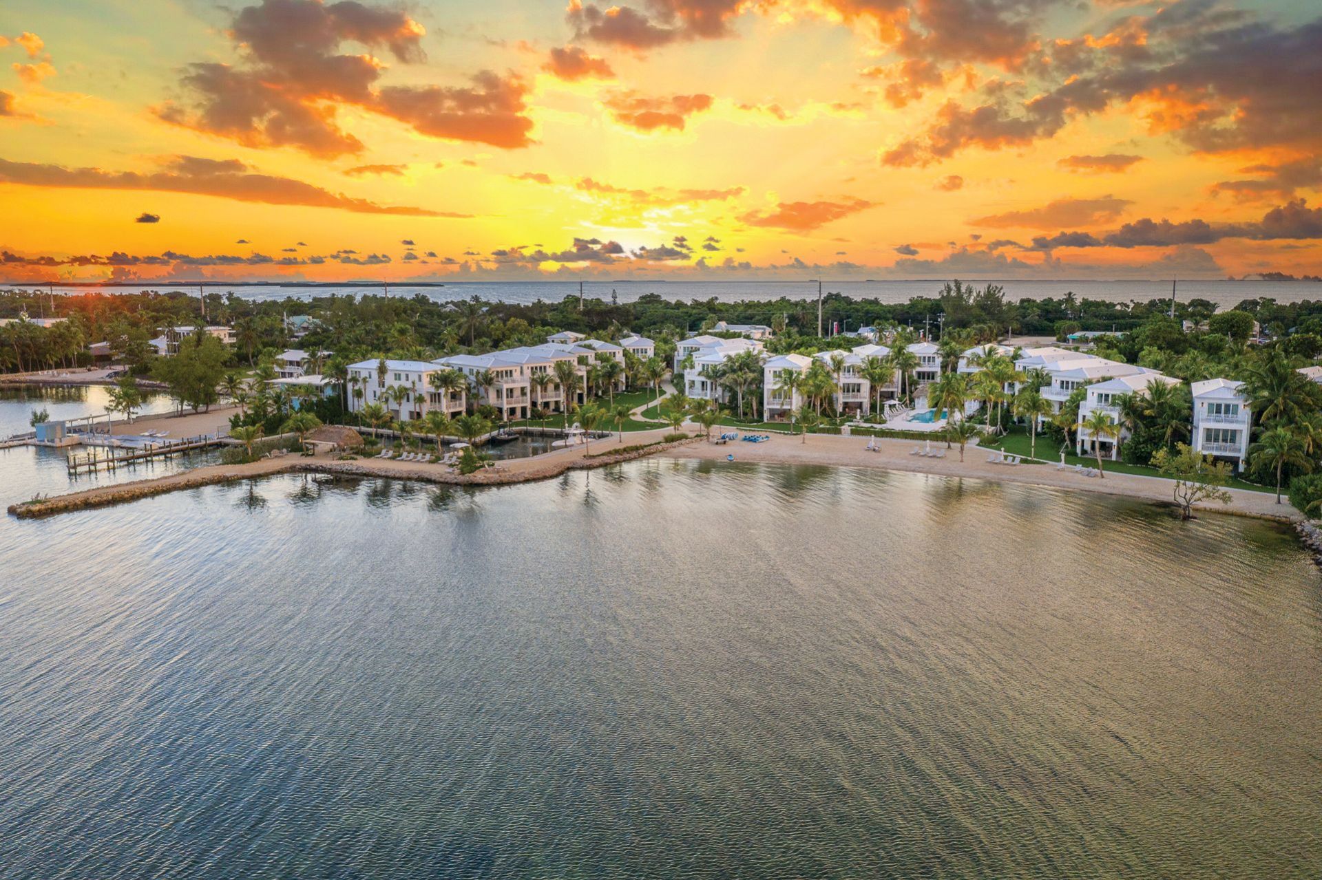 A coastal scene at sunset, with buildings and greenery near the shoreline under a dramatic sky with orange and yellow hues.