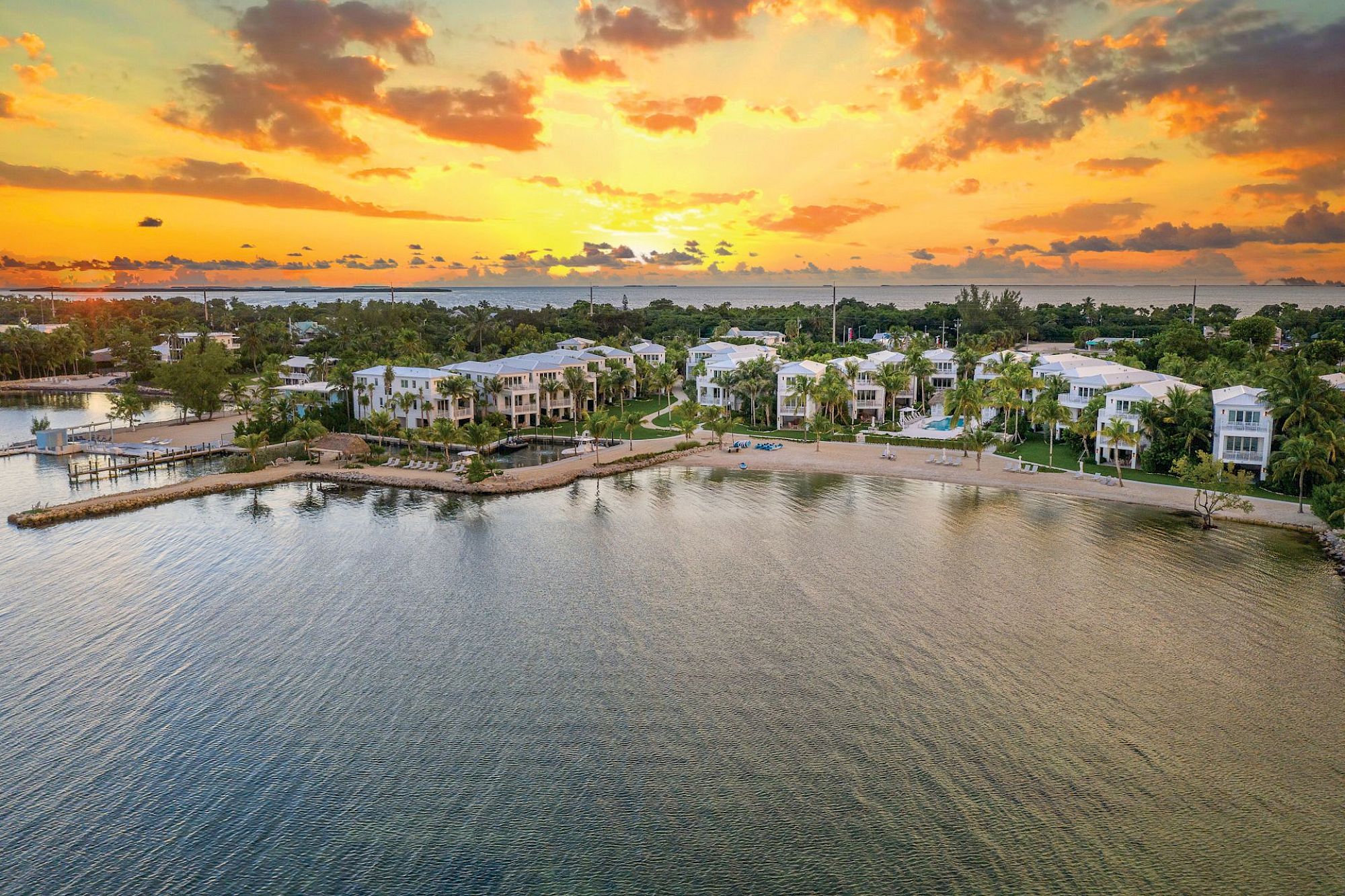 A coastal scene at sunset, with buildings and greenery near the shoreline under a dramatic sky with orange and yellow hues.