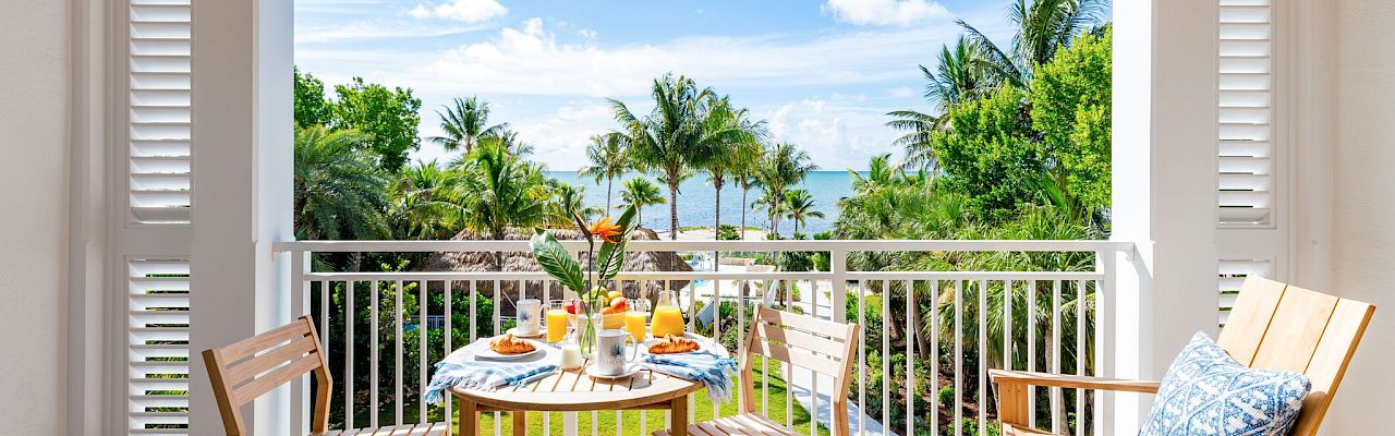 A balcony with a table set for breakfast, surrounded by wooden chairs, overlooks a lush garden and ocean view under a sunny sky.