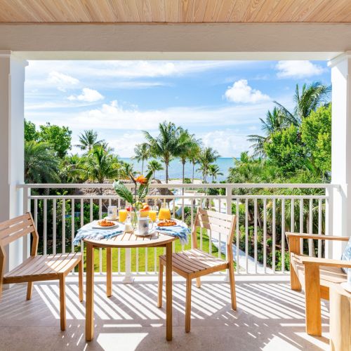 A balcony with a table set for breakfast, surrounded by wooden chairs, overlooks a lush garden and ocean view under a sunny sky.