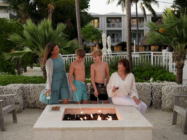 A group of people gather around a fire pit outdoors, surrounded by palm trees and a building in the background.