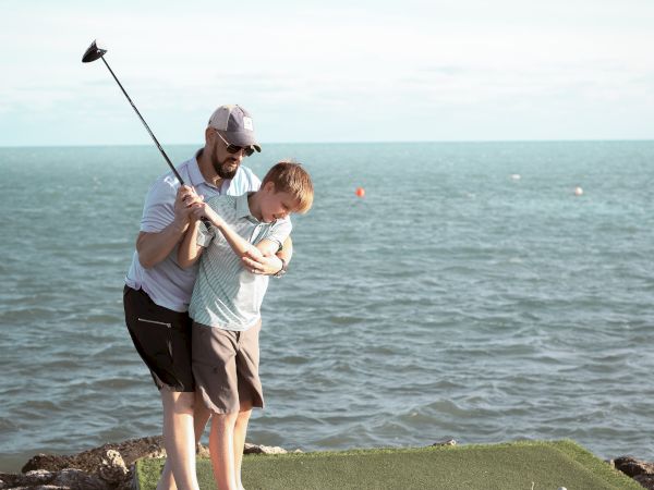 A person guides a child in swinging a golf club on a grass platform by the sea, under a clear sky.