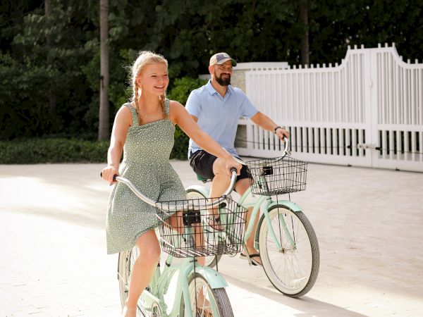 Two people are riding bicycles on a sunny path, surrounded by trees and a white fence.