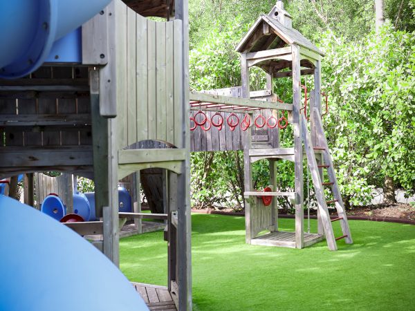 The image shows a playground with a blue slide, wooden climbing structures, and green artificial turf, surrounded by trees.