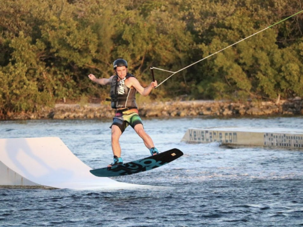 A person is wakeboarding on water, holding a rope, wearing a life jacket and helmet, and doing a jump near a ramp.