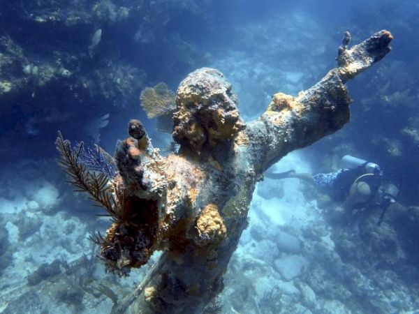 A coral-encrusted underwater statue with outstretched arms, accompanied by a scuba diver exploring the ocean surroundings.